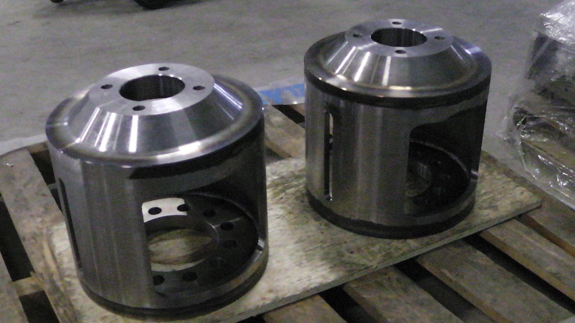 Deux composants cylindriques en métal usiné, présentant de larges découpes latérales rectangulaires, reposent sur une palette en bois dans un atelier.