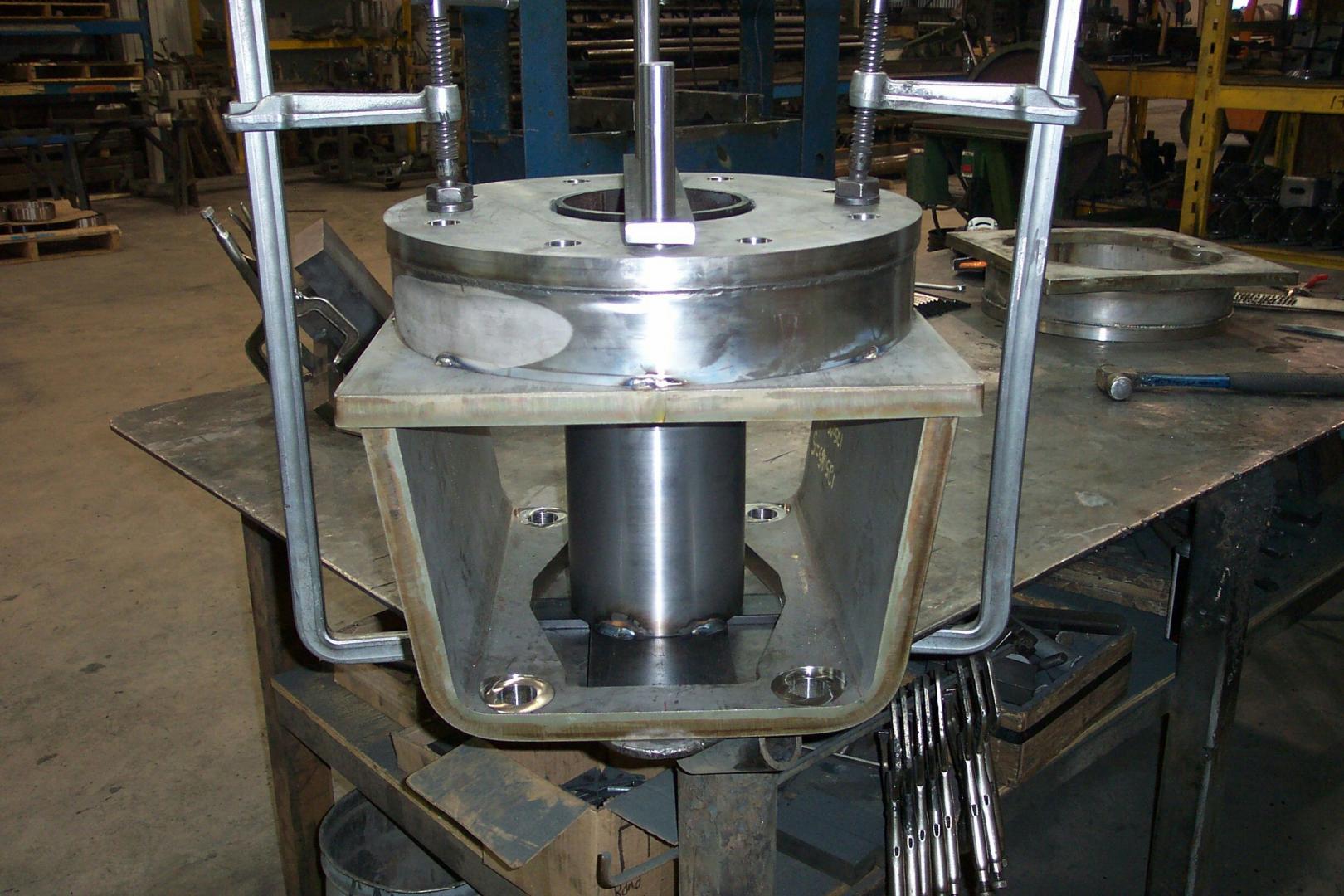 Two metal clamps hold a cylindrical metal component to a steel bracket on a workbench in a machine shop.