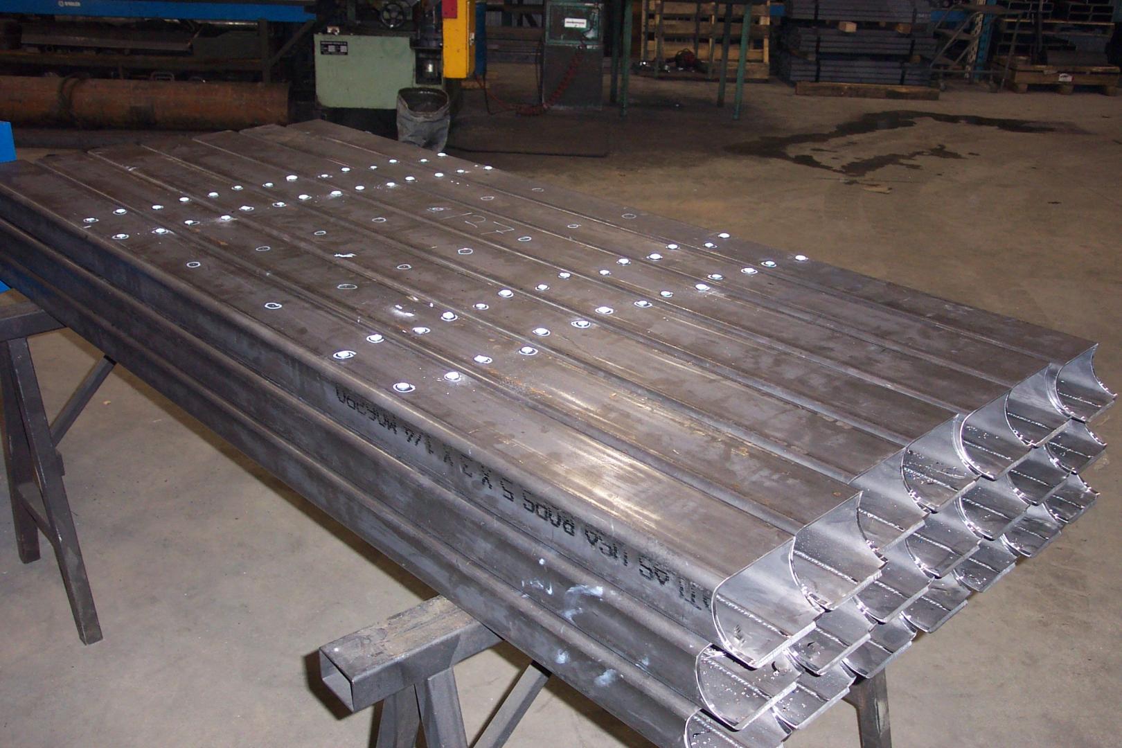 A stack of long, metal channel bars with pre-drilled holes resting on two metal sawhorses in a workshop setting.