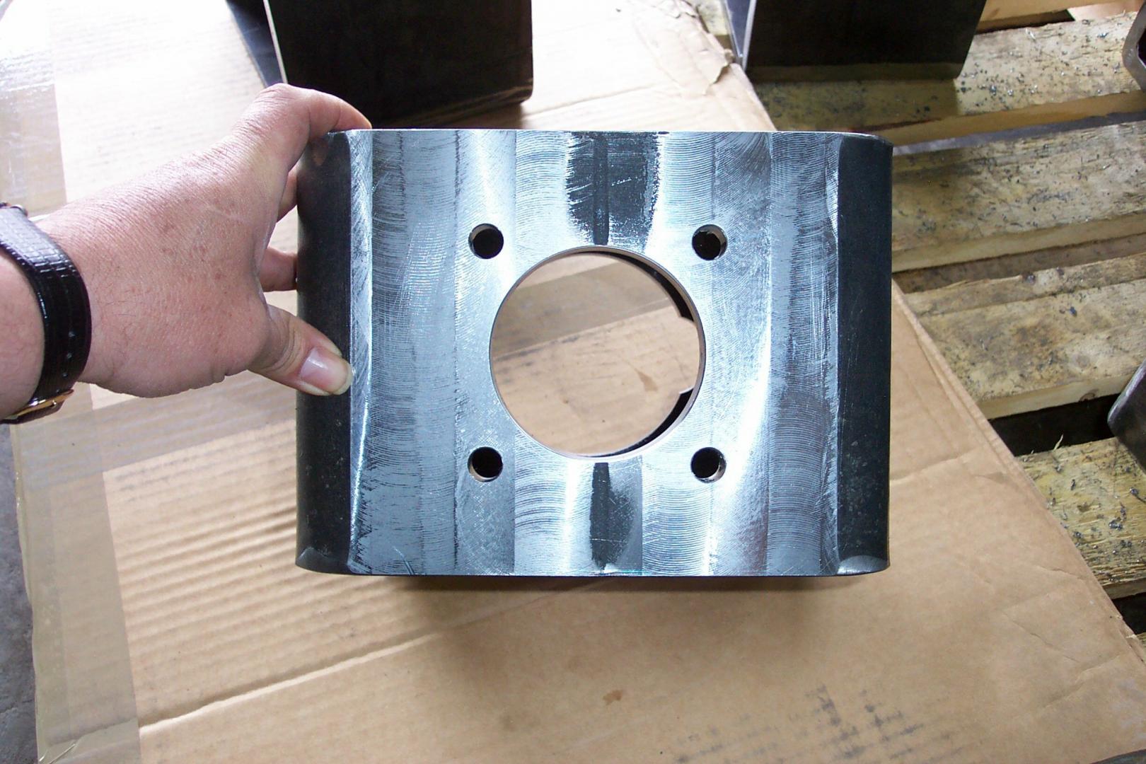 Une main tient un bloc de métal rectangulaire et sombre, percé d'un trou circulaire central et de quatre trous de boulons circulaires plus petits.