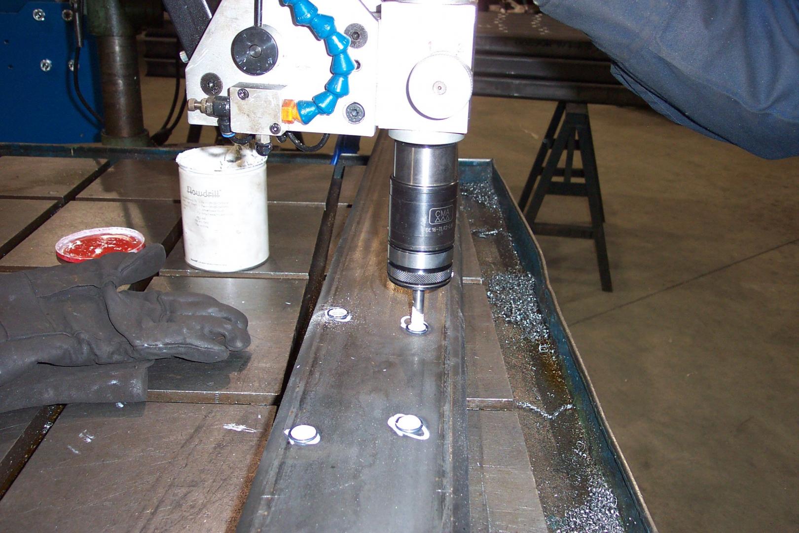 A metal drill press actively boring holes into a flat steel beam in a workshop setting.