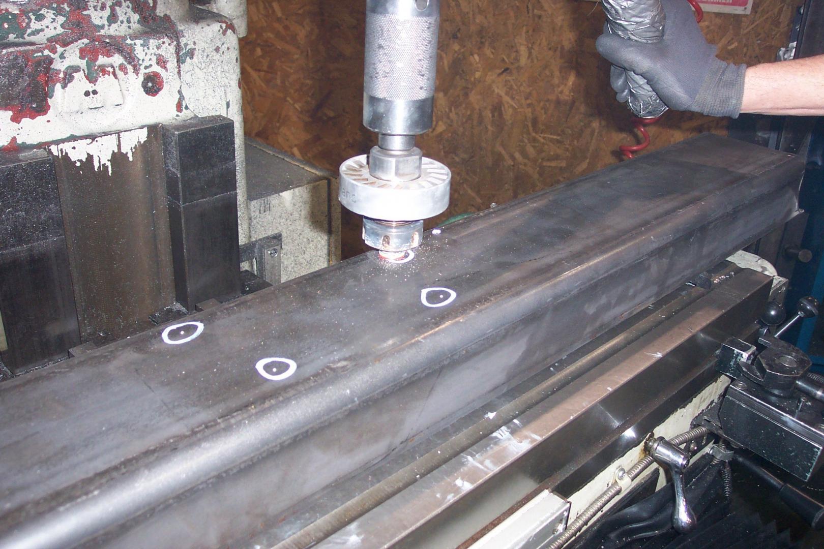 A machine tool uses a drill bit to create holes in a long, dark metal bar marked with white circles.
