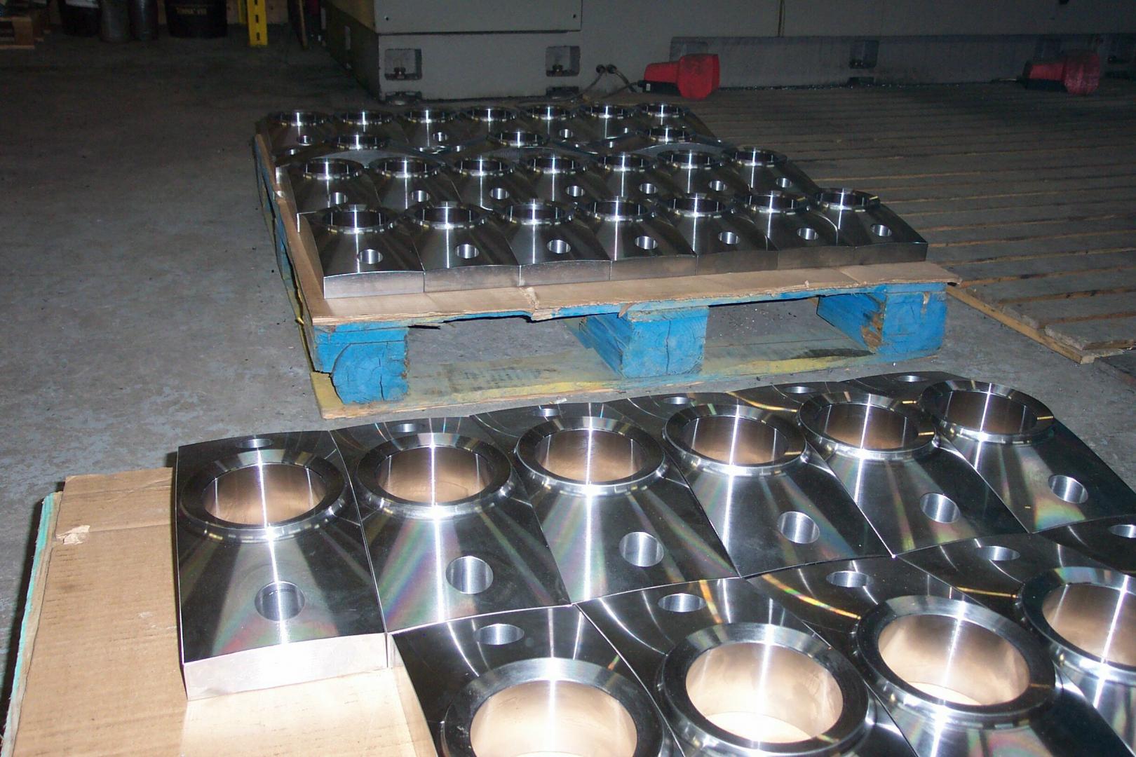 Des rangées de pièces industrielles métalliques usinées, comportant des ouvertures circulaires, sont empilées sur des palettes en bois dans un atelier.
