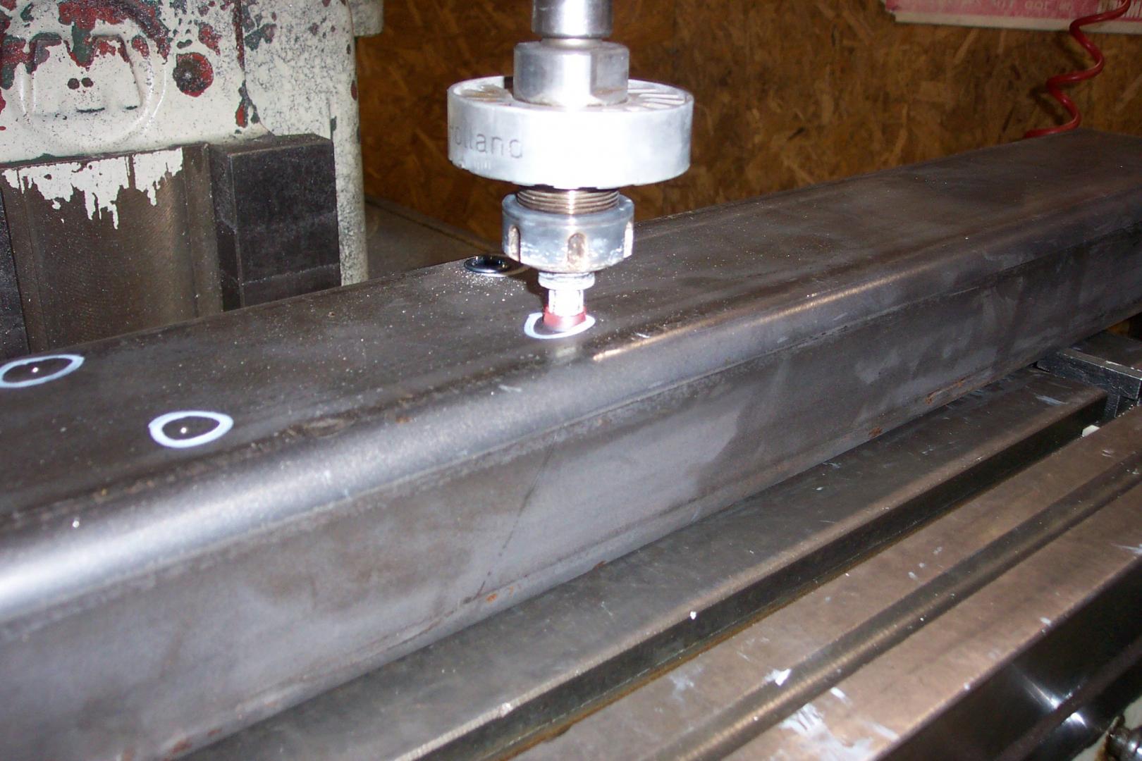 A metal milling machine drills into a steel beam marked with white circles for hole locations.