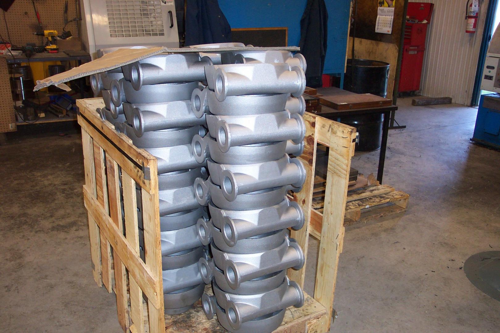 Deux piles de pièces industrielles cylindriques identiques en métal gris, stockées dans une caisse en bois à l'intérieur d'un atelier.