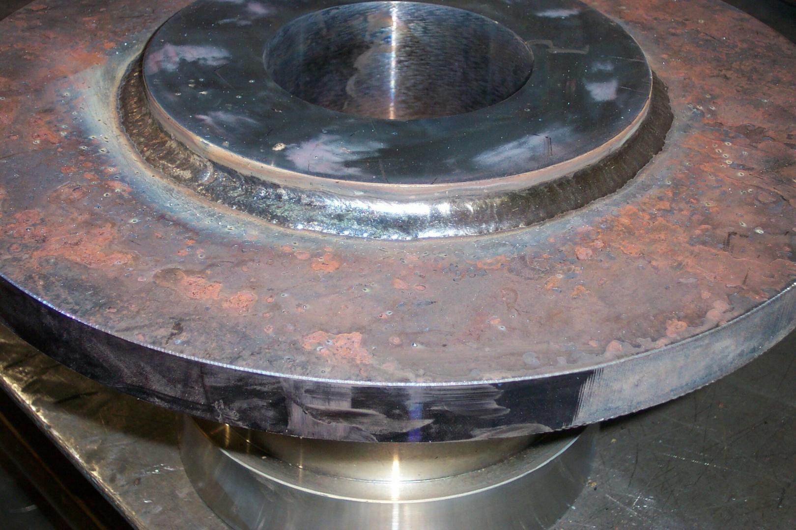 A metal pipe flange welded to a circular plate, showing a clean, shiny weld bead amidst rusted, unpainted steel surfaces.