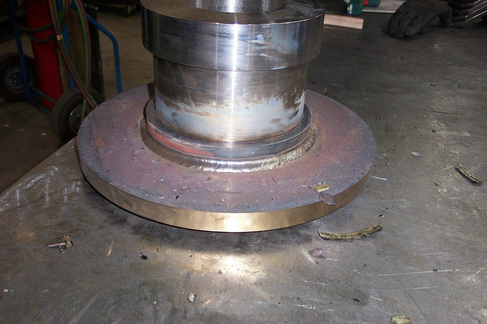 A metallic, cylindrical shaft welded to a flat circular plate sits on a workshop workbench.