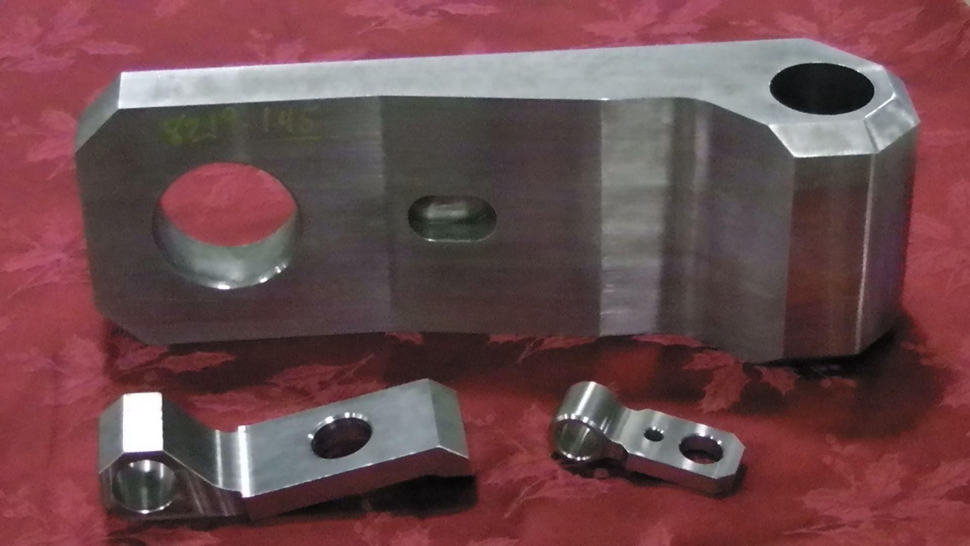 Trois pièces industrielles en métal argenté usiné, de tailles différentes, disposées sur une surface à motifs rouges.