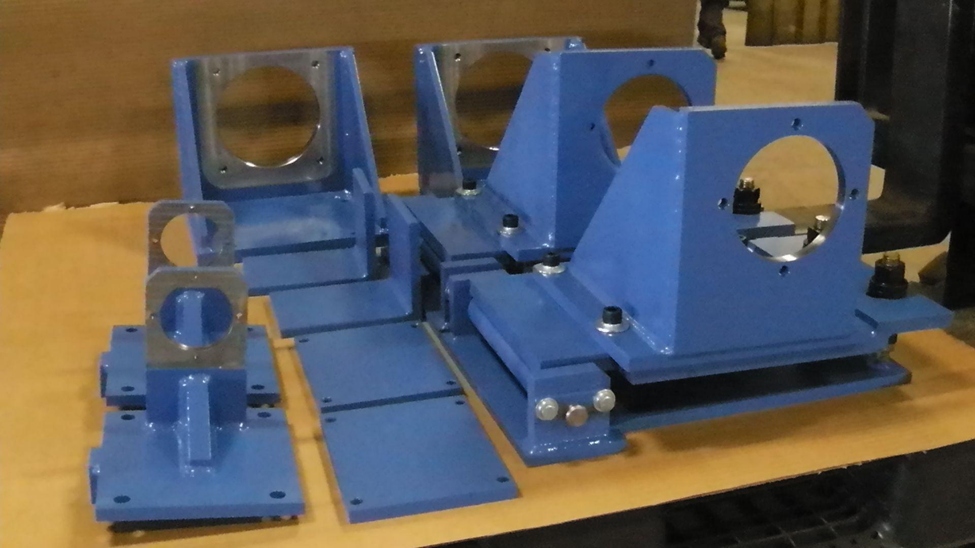 Several blue, L-shaped industrial metal mounts with circular faceplates sit on a flat, cardboard-covered surface.