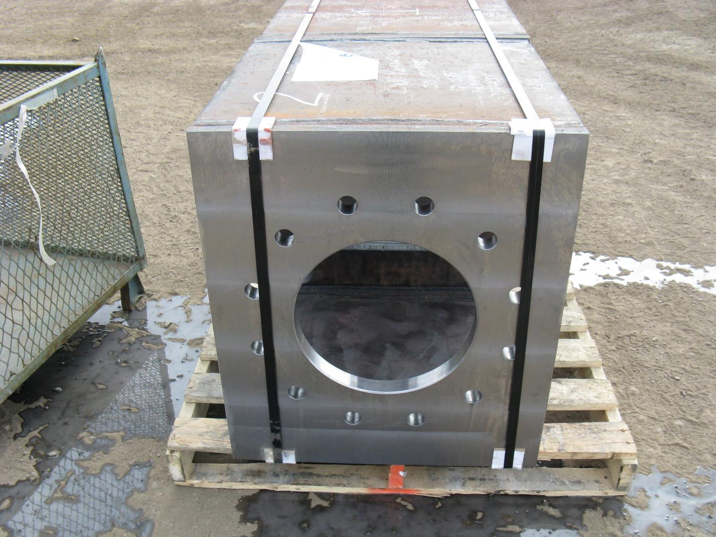 Un gros bloc de métal gris rectangulaire, percé d'une ouverture circulaire centrale et de trous pour boulons, est fixé sur une palette en bois.