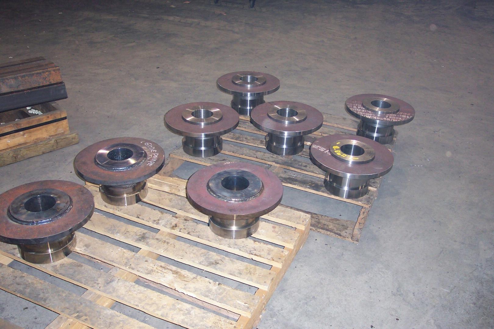 Eight metallic, flanged industrial hubs are arranged on a wooden shipping pallet in an industrial facility.