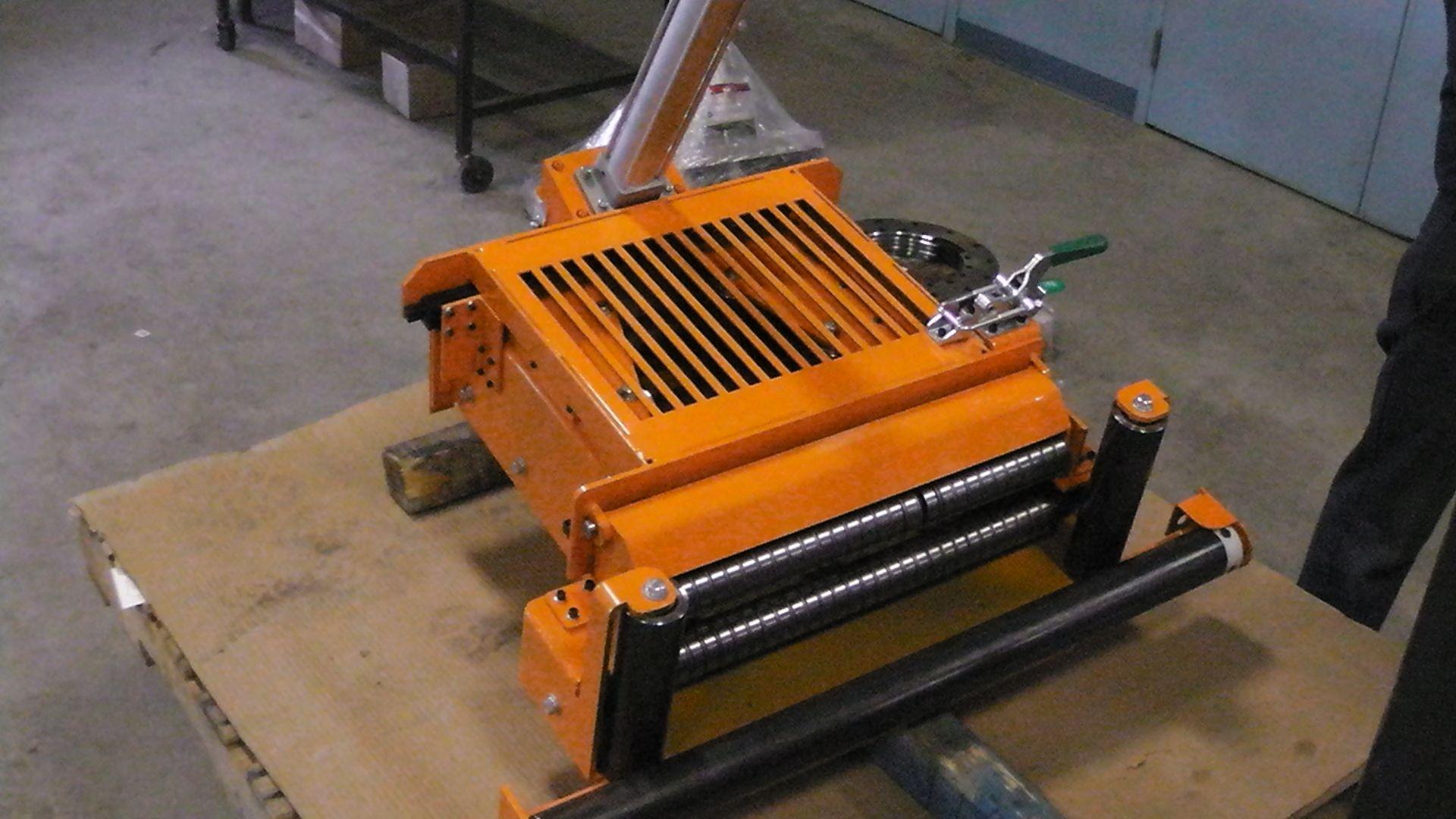An orange, industrial metal machine with rollers and a grill sits on a wooden pallet in a workshop.