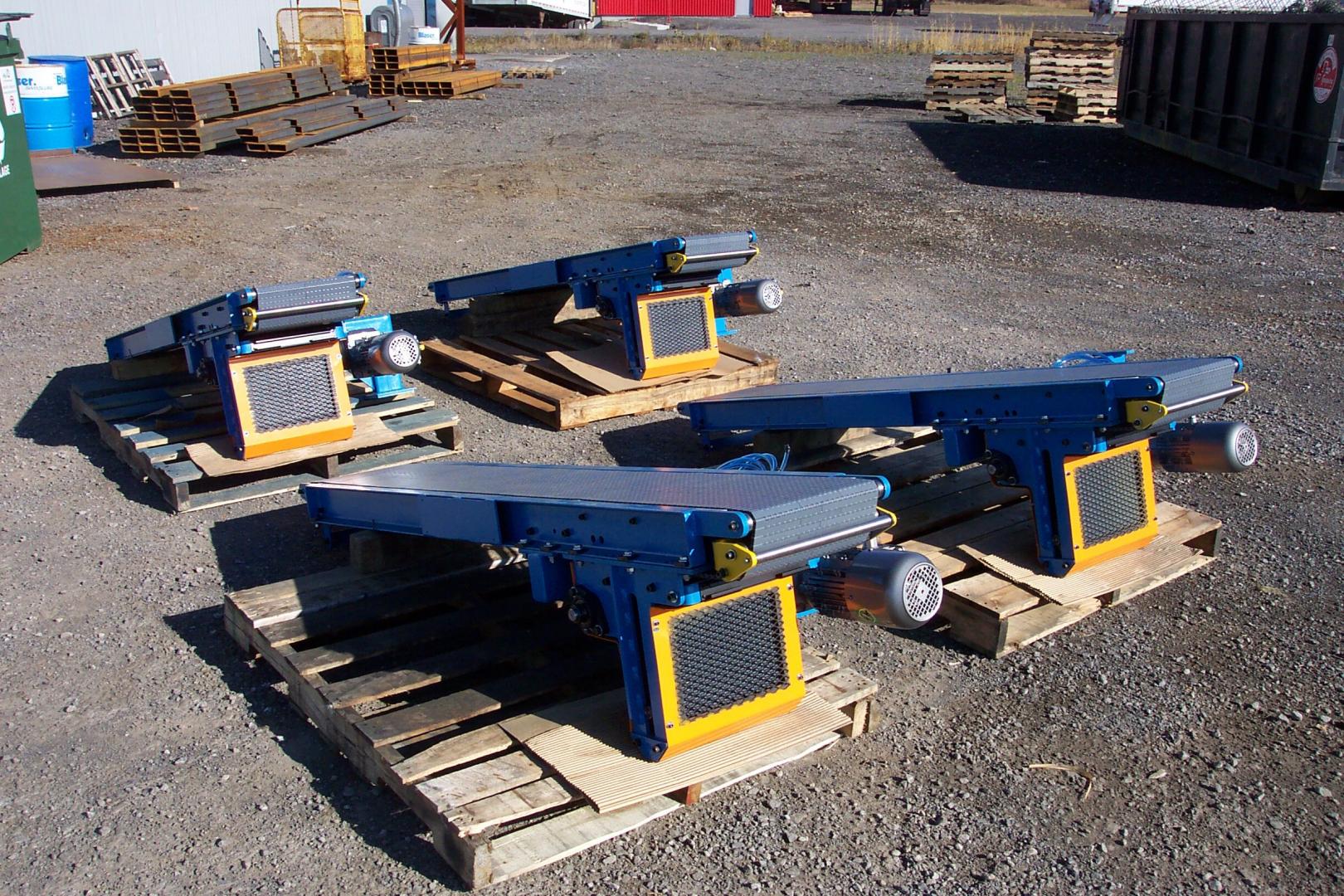 Four blue and yellow industrial conveyor units on wooden pallets in an outdoor gravel lot.