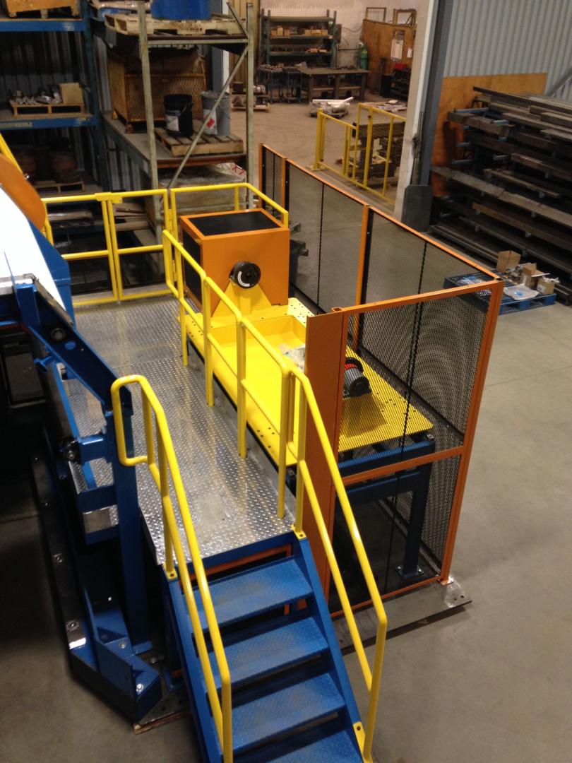 A yellow industrial machine sits on a raised metal platform with blue stairs and safety railings in a warehouse.