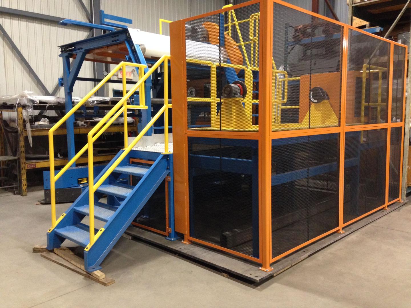 Industrial machinery on a platform with blue stairs and yellow railings inside a warehouse enclosed by a metal cage.