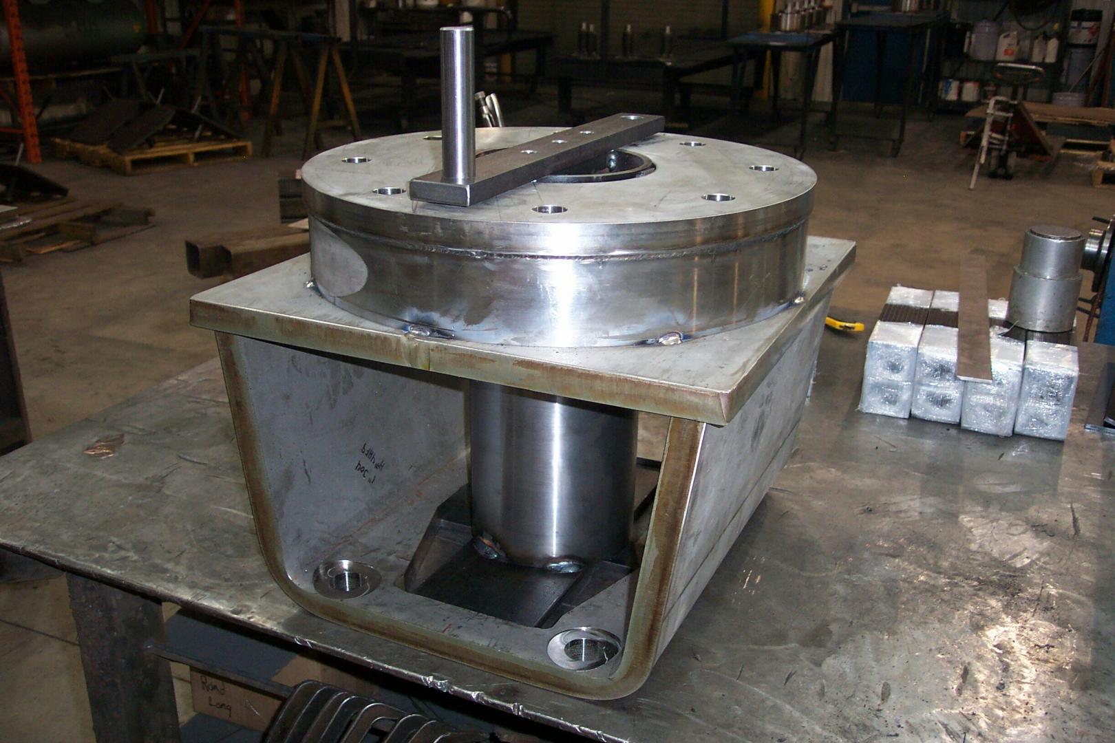 A metal hydraulic cylinder assembly sits inside a U-shaped steel frame on a workshop table.
