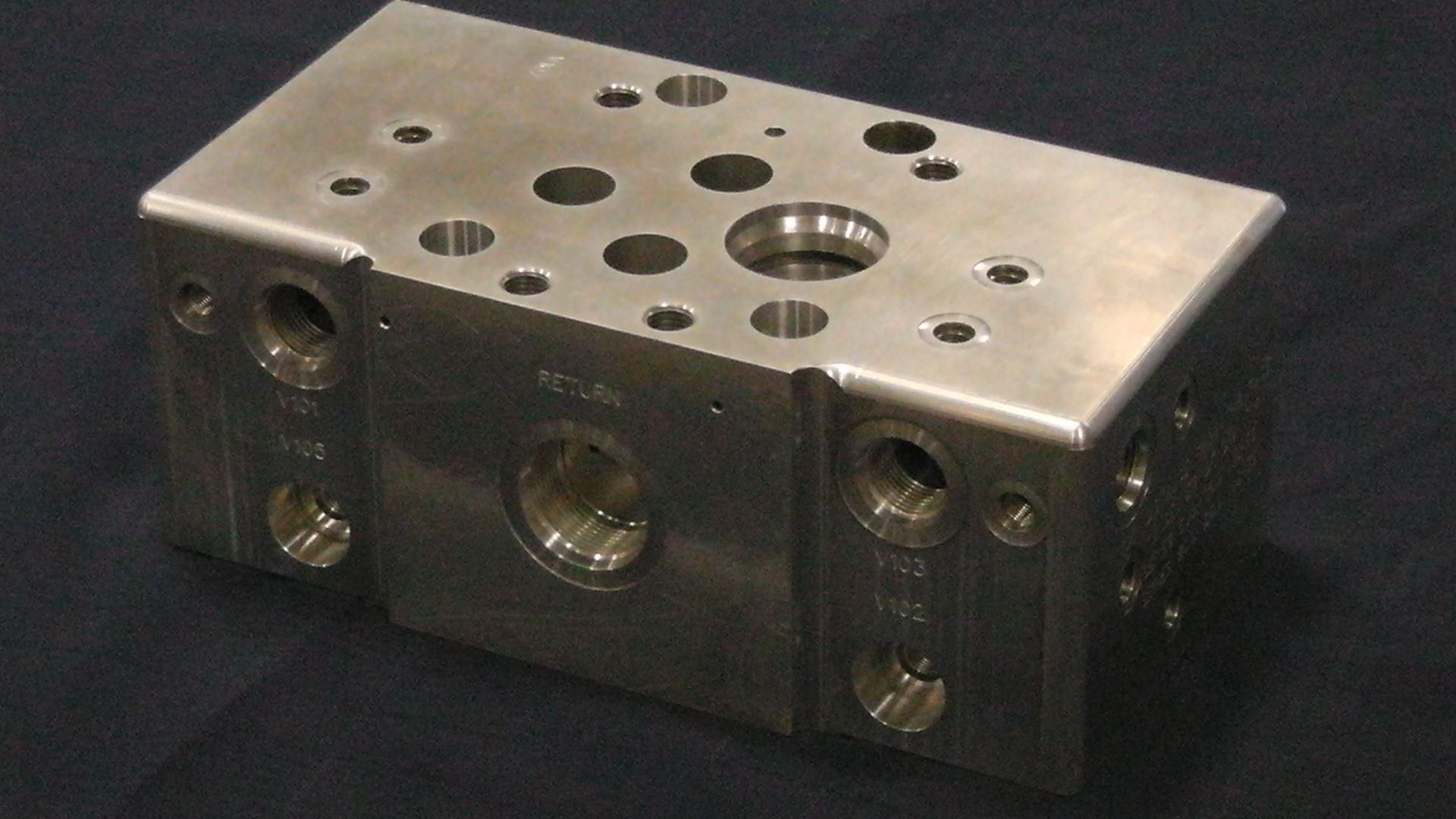 Un bloc collecteur hydraulique rectangulaire en métal avec de multiples orifices percés et trous de fixation, sur fond noir.