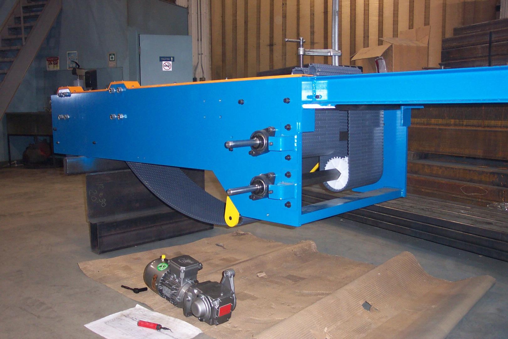 A blue machinery assembly sits in a workshop with a motor on the floor and a screwdriver resting on a nearby paper.