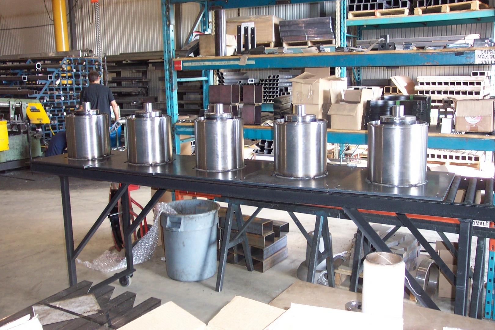 Five identical metal industrial cylinders stand in a row on a black workbench inside a manufacturing facility.