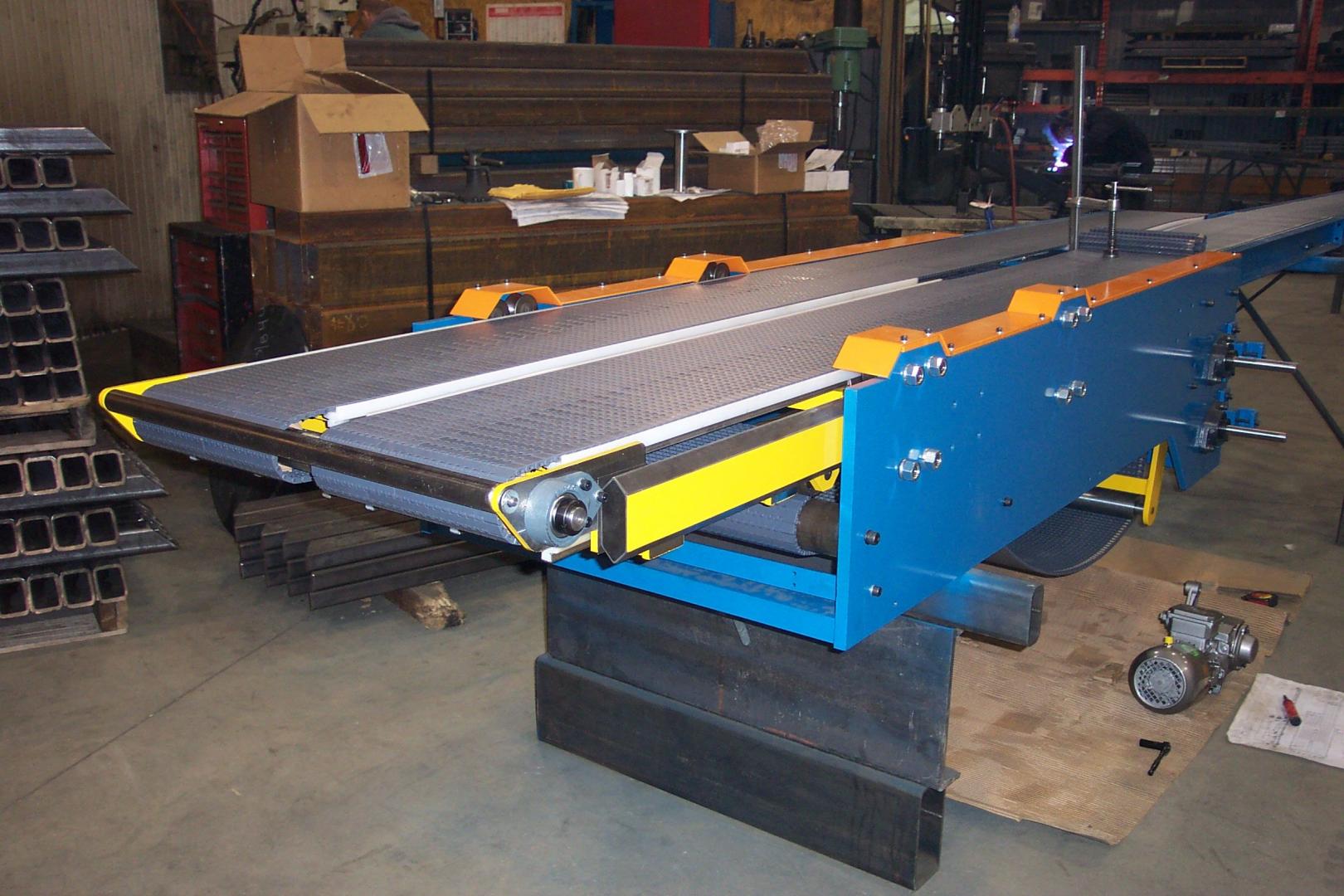 A blue industrial belt conveyor with yellow safety guards and a textured surface sits in a workshop setting.