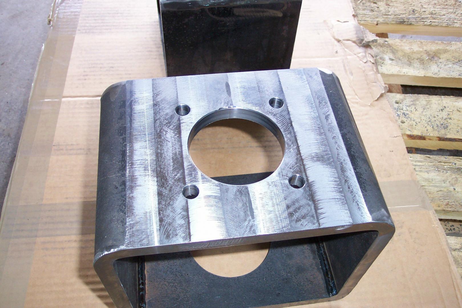 Un support de montage carré en métal creux, avec un trou circulaire central et quatre trous de boulon, sur une surface en carton brun.