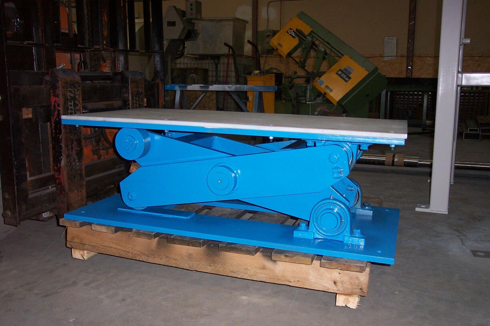 A bright blue industrial scissor lift table sits on a wooden pallet in a workshop setting.