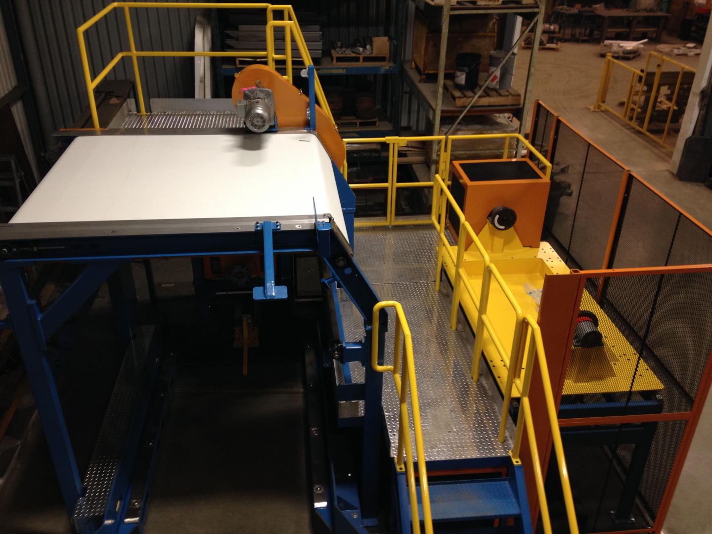 An industrial workstation with blue and yellow metal platforms, stairs, safety railings, and mesh fencing in a warehouse.