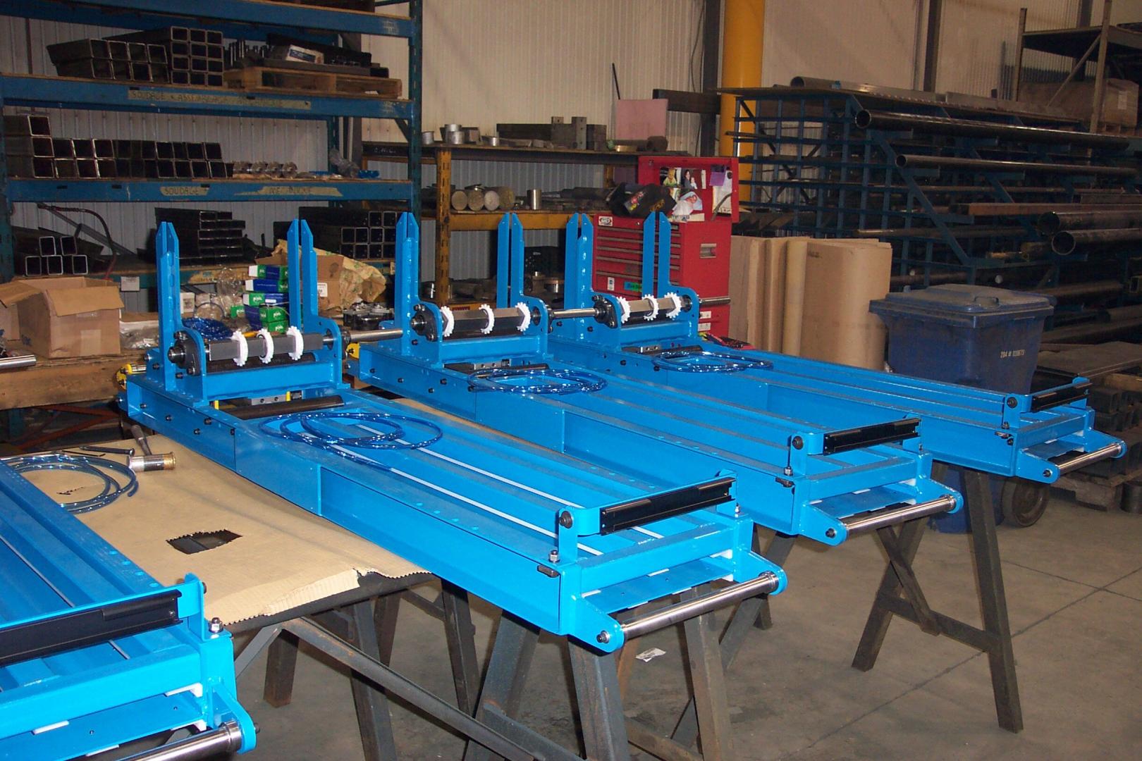 Three bright blue industrial metal lifts sit on sawhorses inside a warehouse workshop with storage racks in the background.