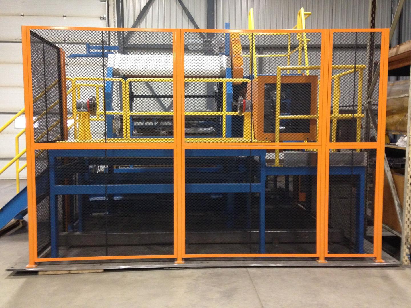 Industrial machinery enclosed by an orange metal safety cage with black wire mesh, set inside a warehouse facility.