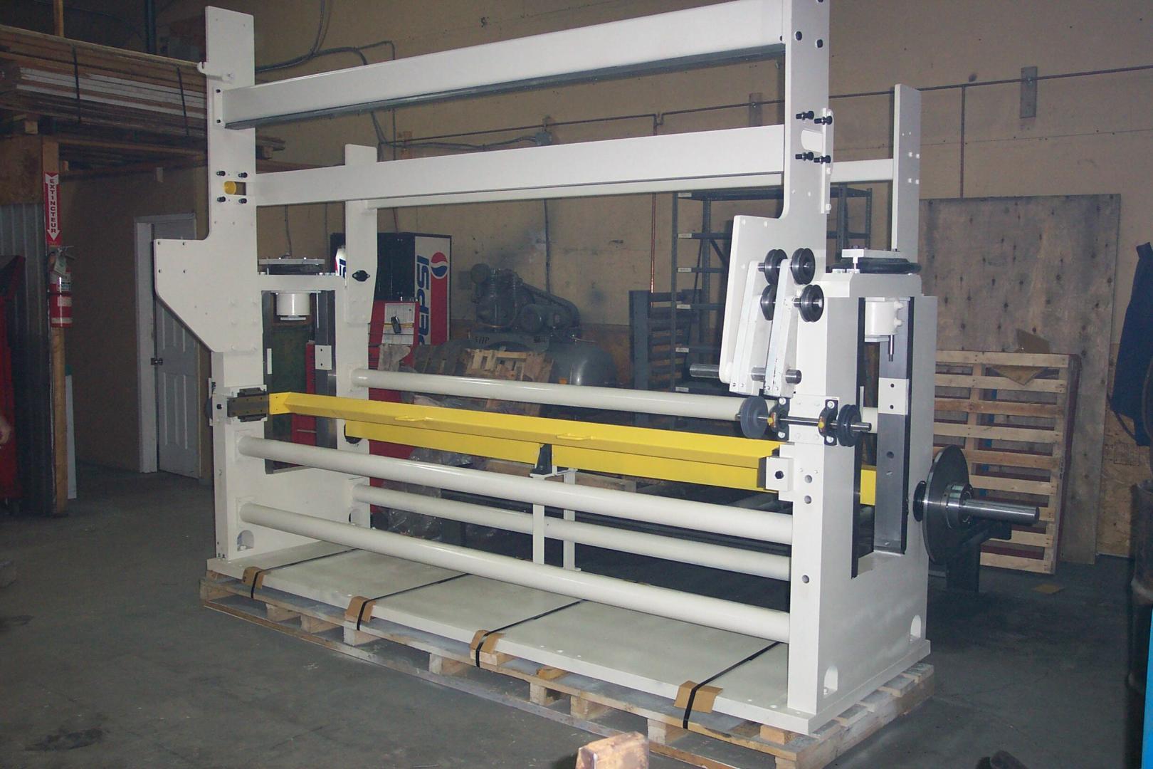 A large, white industrial machinery frame with a prominent yellow horizontal bar, sitting on wooden pallets in a workshop.