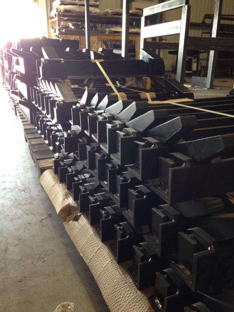 A large stack of identical black metal industrial parts arranged in rows on a pallet in a warehouse setting.