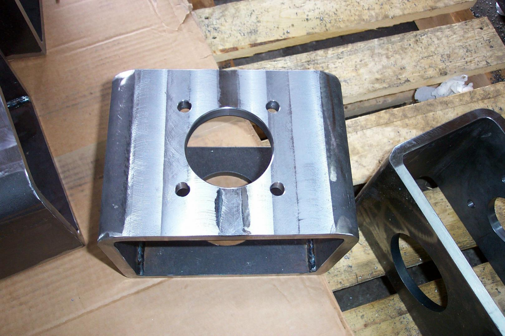 Une équerre métallique rectangulaire avec un trou central circulaire et quatre petits trous de boulons, fixée sur une palette en bois.