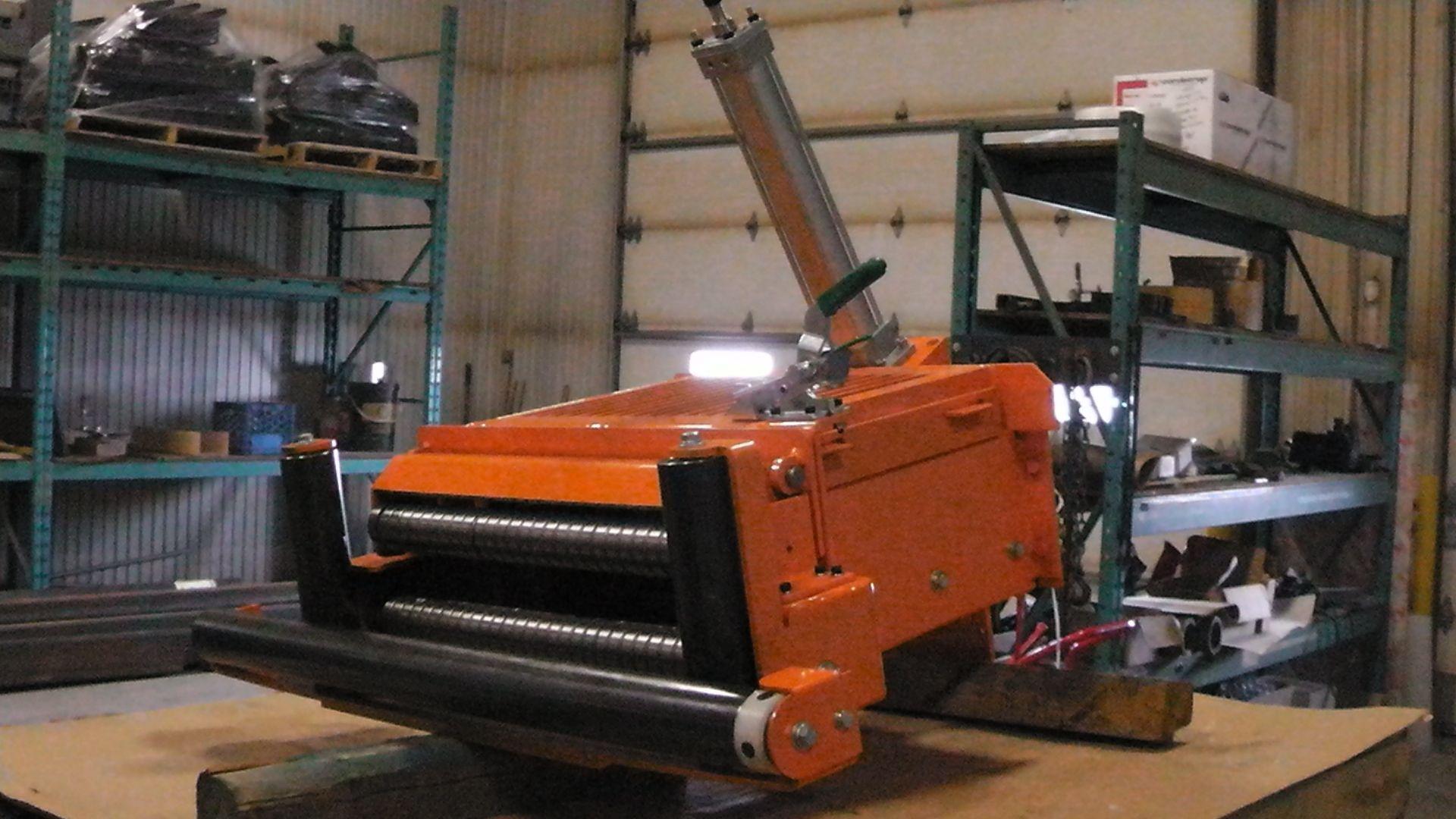 An orange industrial metal-feeding machine with rollers, sitting on a workbench in a storage warehouse.