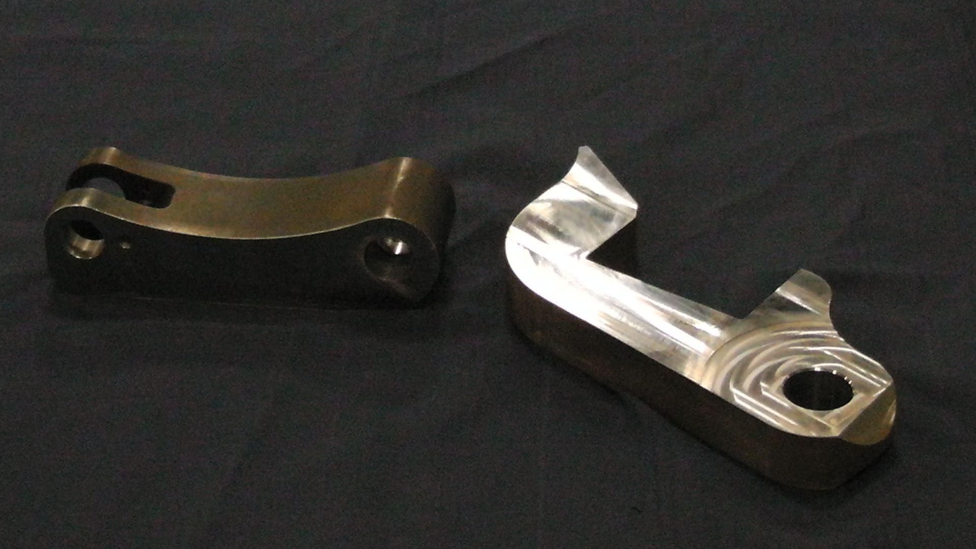 Deux pièces mécaniques métalliques, percées de trous et de formes courbes, reposent sur une surface en tissu sombre.