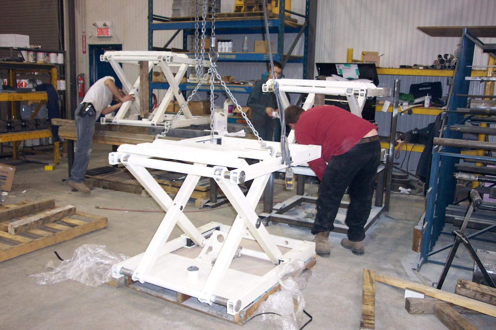 Workers assemble large, white metal scissor lift mechanisms in a brightly lit industrial workshop.
