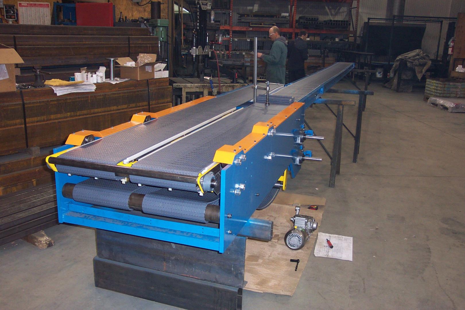 A blue and orange industrial conveyor machine sits in a workshop, with a person working in the background.