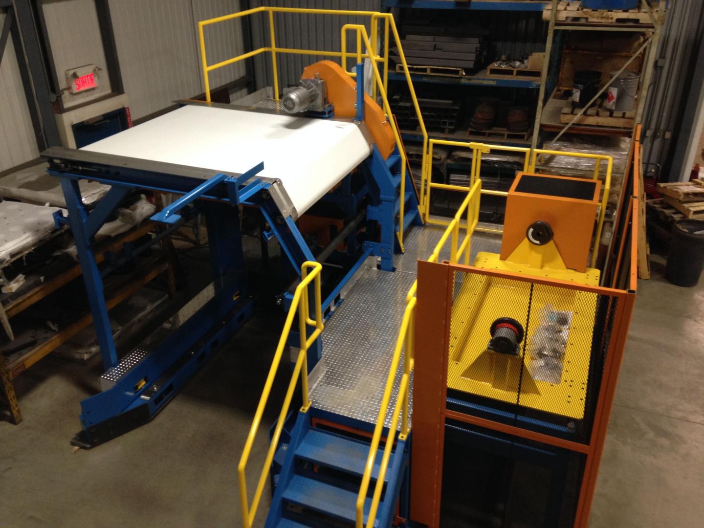 Industrial machinery featuring blue framing, a white conveyor belt, and yellow safety railings in a warehouse setting.