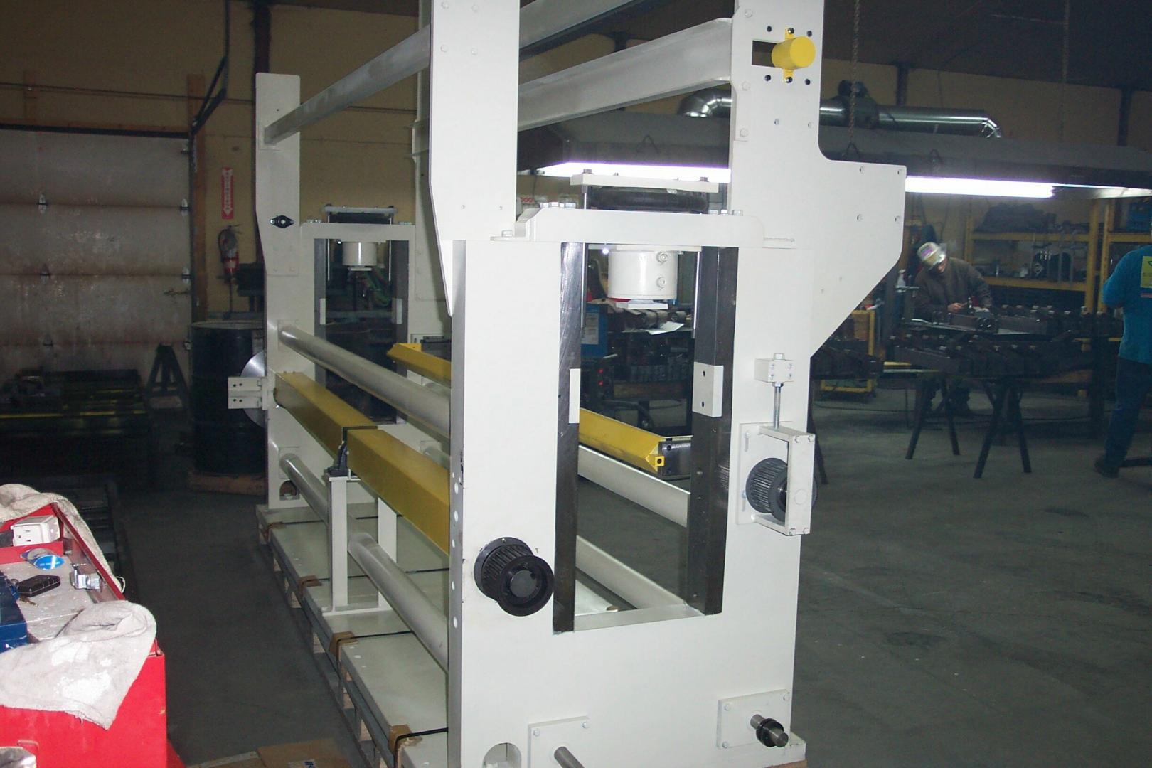 A large, white industrial metal frame machine stands in a workshop setting with yellow rollers and mechanical components.