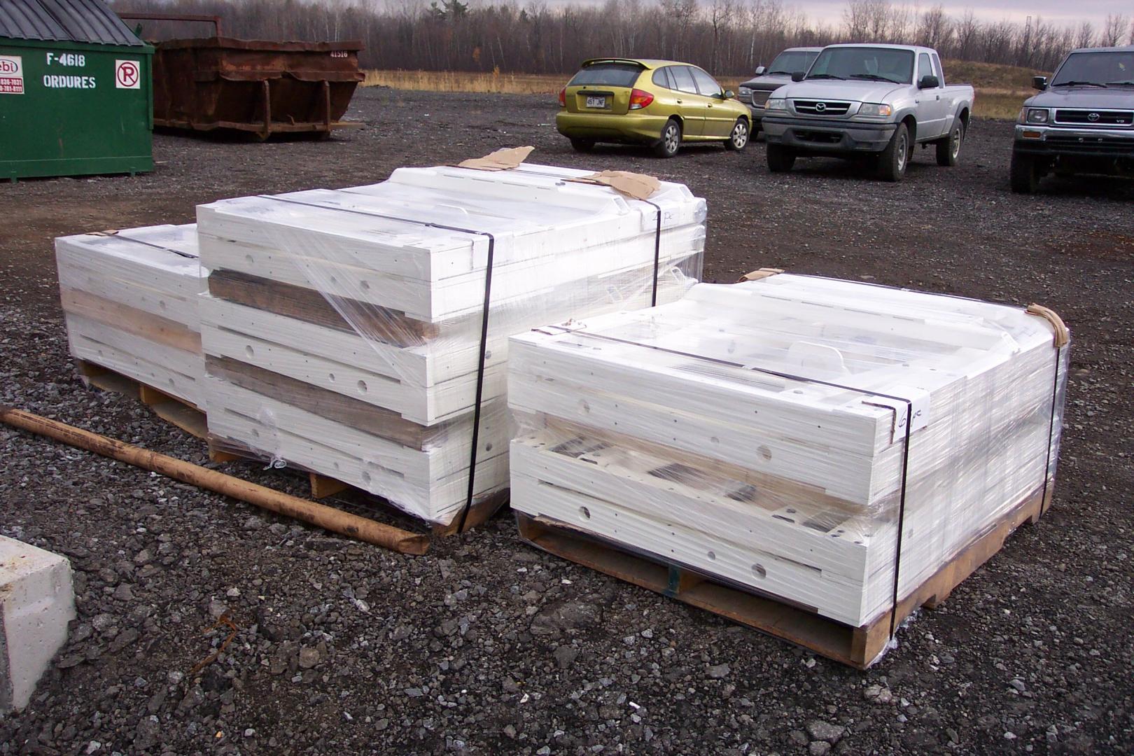 Three pallets of stacked white, shrink-wrapped construction panels sitting on a gravel lot near several parked vehicles.