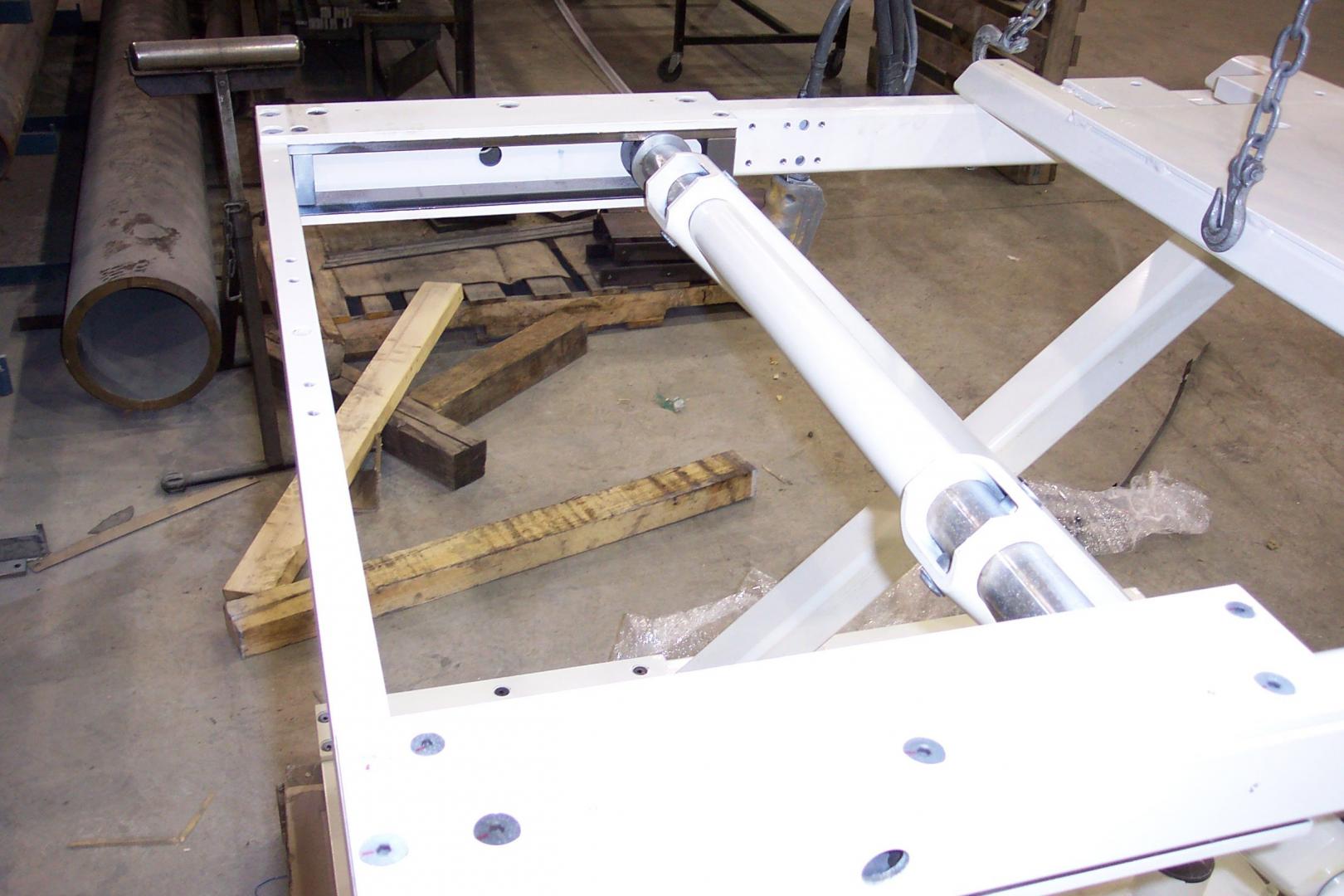 White metal frame assembly featuring a hydraulic cylinder mounted between beams in an industrial workshop.