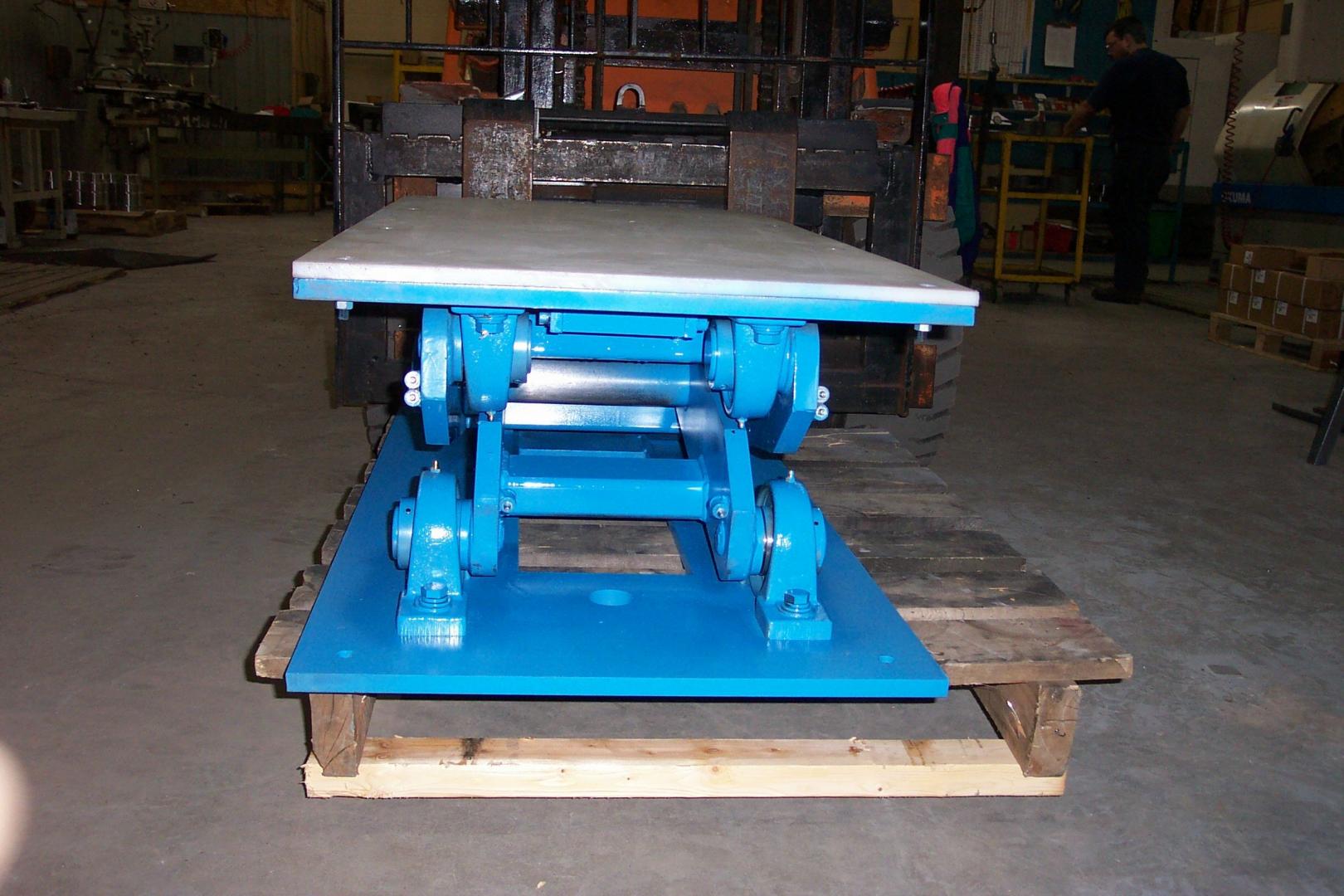 A blue metal hydraulic scissor lift table sits on a wooden pallet inside a workshop.