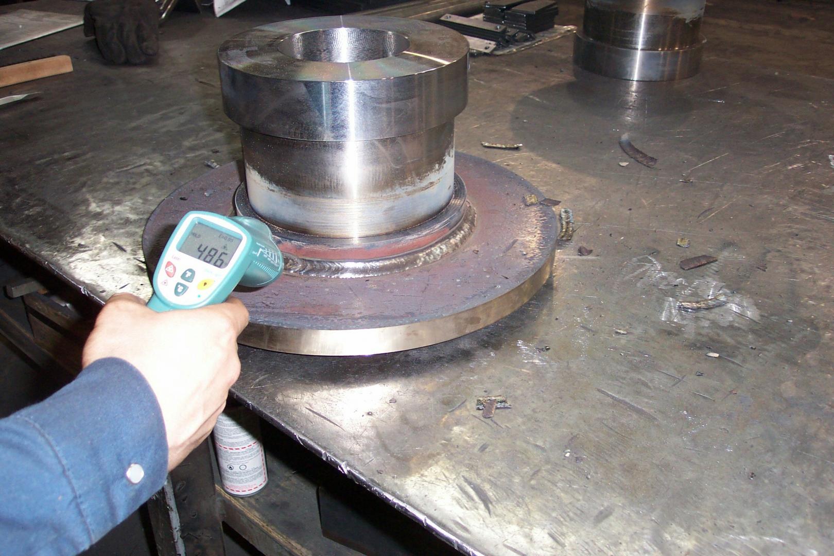 A hand uses a digital infrared thermometer to measure the temperature of a welded metal component on a workshop table.