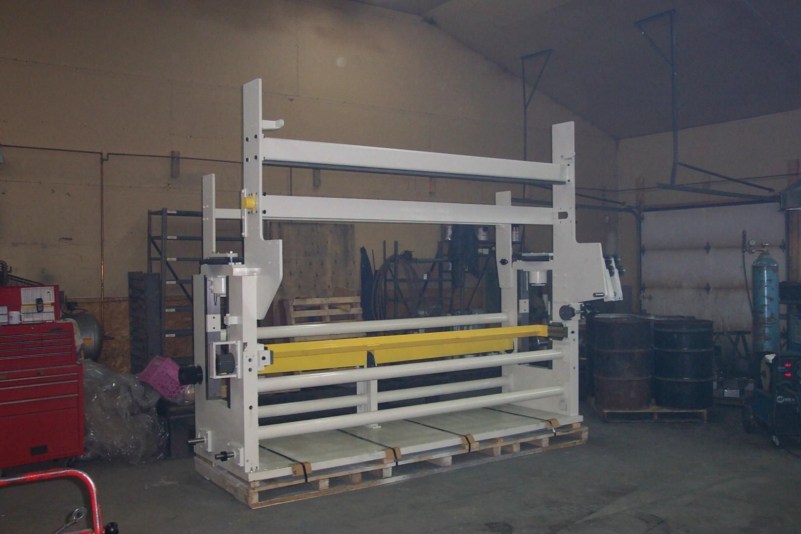 A large, white industrial machinery frame with a yellow roller sits on a pallet in a warehouse workshop.