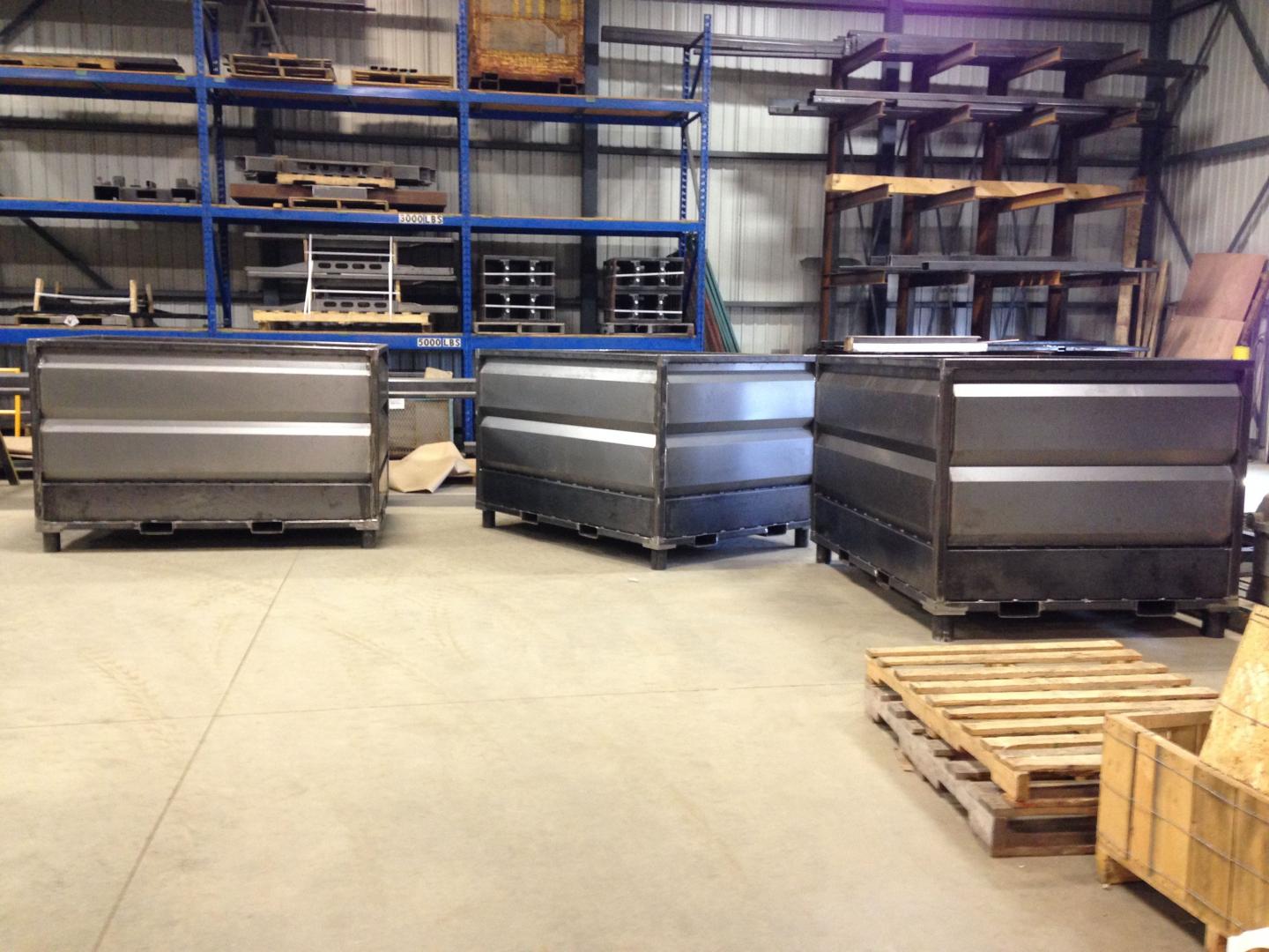 Three large, grey metal industrial bins stand on a warehouse floor in front of tall shelving units.
