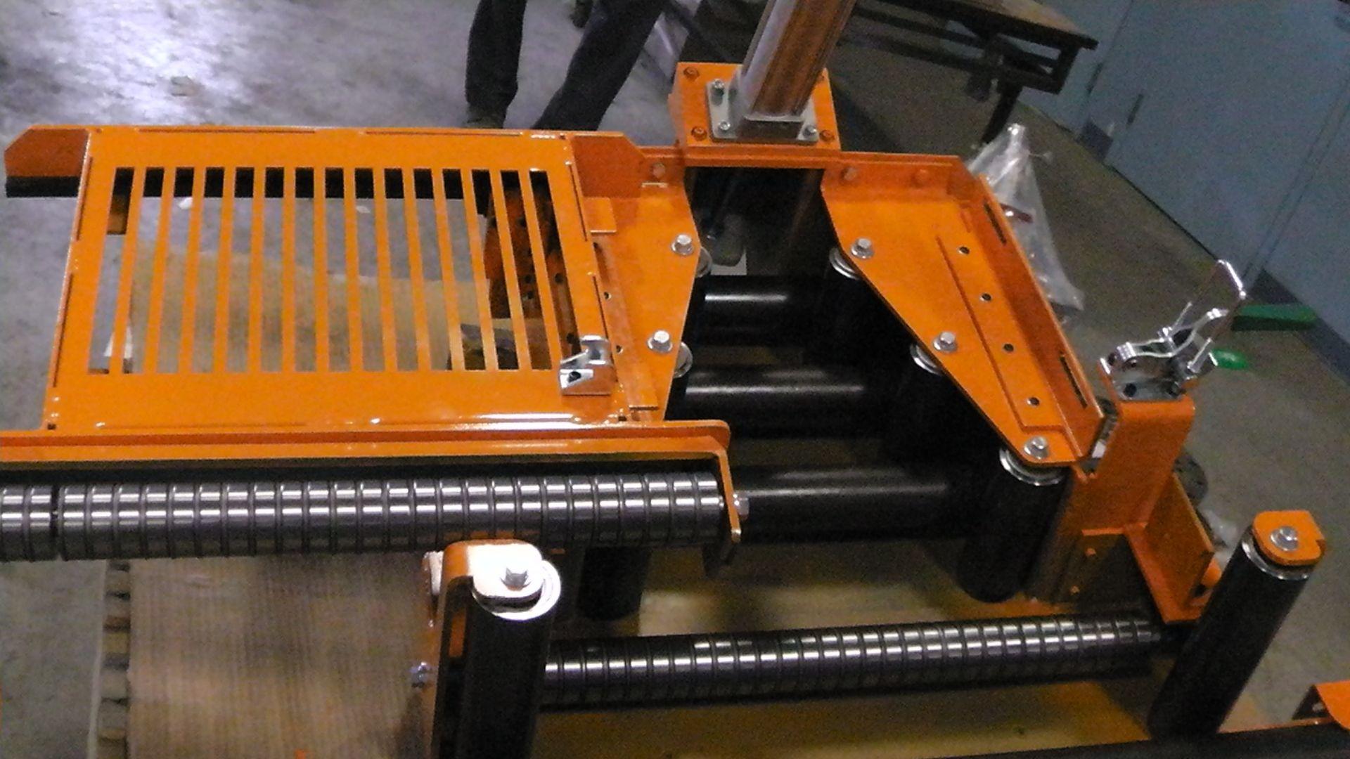 A mechanical roller assembly with bright orange metal housing and black cylindrical rollers on a shop floor.