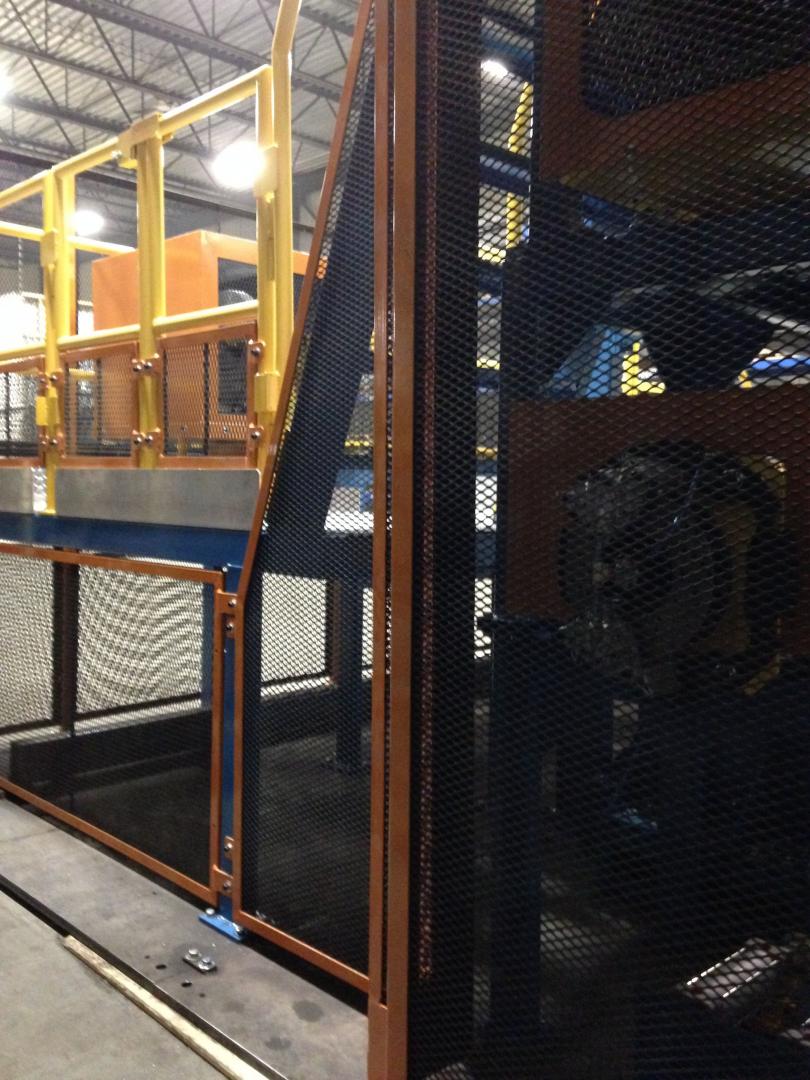 Industrial machinery in a warehouse, featuring yellow railings and a black metal safety mesh enclosure.