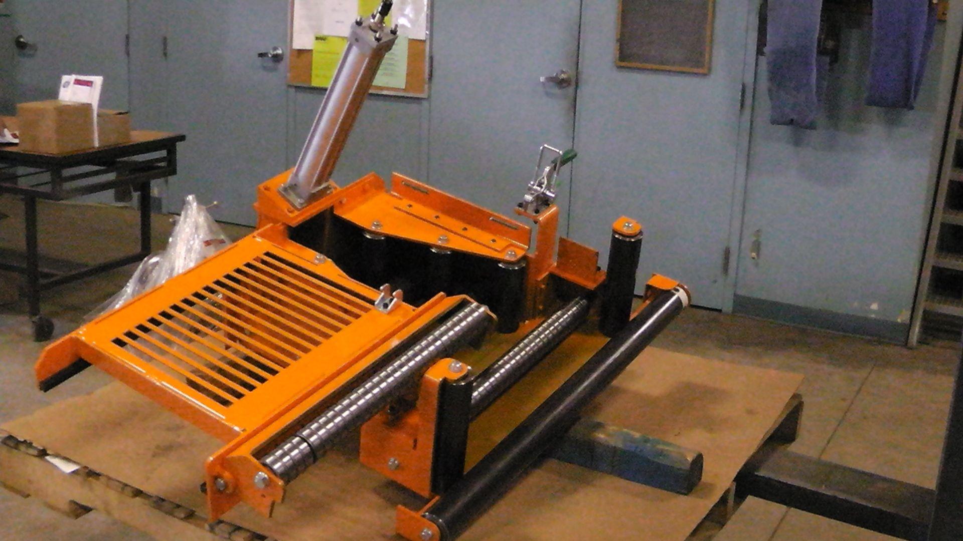 An orange, industrial metal assembly with guide rails and a pneumatic cylinder sitting on a wooden pallet in a workshop.