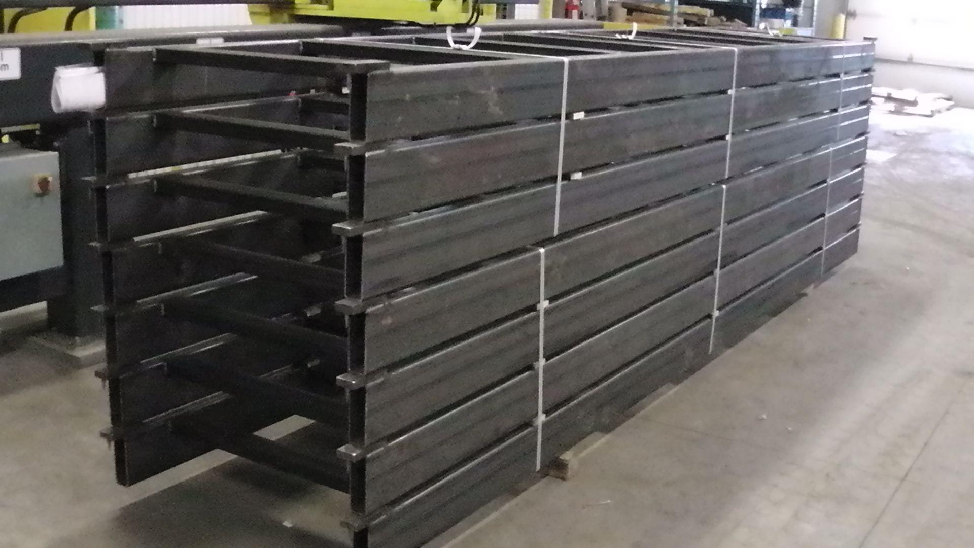 A large stack of long, black metal beams or frames in an industrial workshop setting.