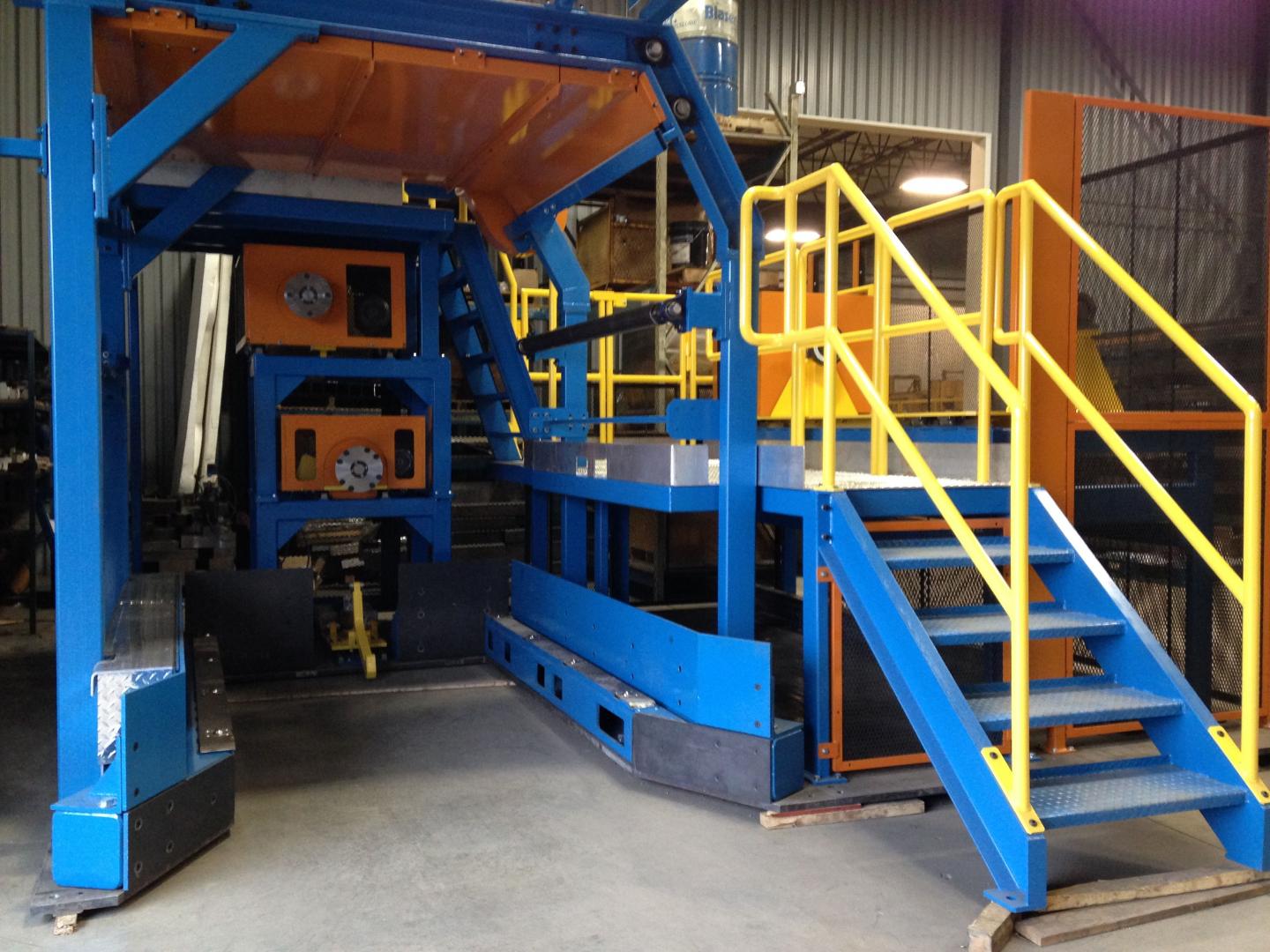 A blue industrial machine with orange accents and a staircase with yellow handrails in a warehouse setting.