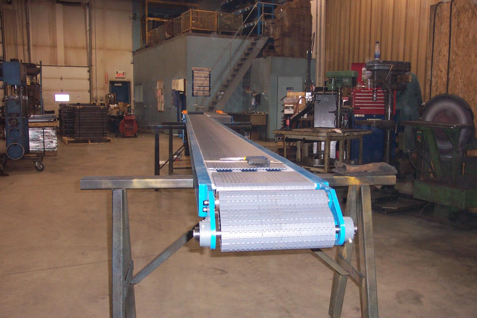 A long, blue-framed industrial conveyor belt on metal stands inside a warehouse shop.